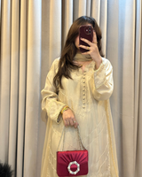 Woman taking a mirror selfie wearing a light-colored traditional outfit with a red handbag against a beige curtain background.