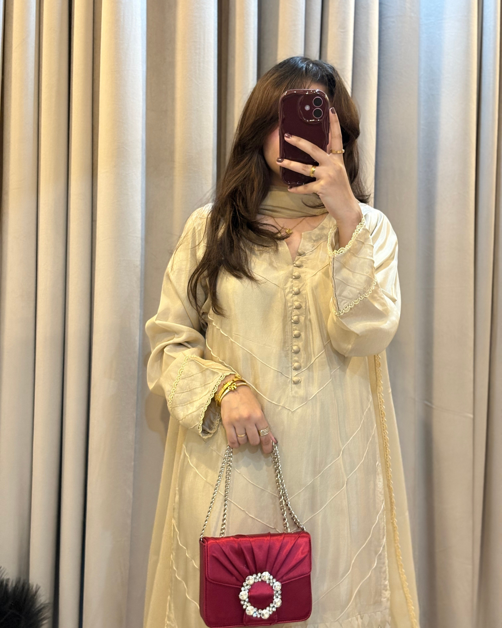 Woman taking a mirror selfie wearing a light-colored traditional outfit with a red handbag against a beige curtain background.
