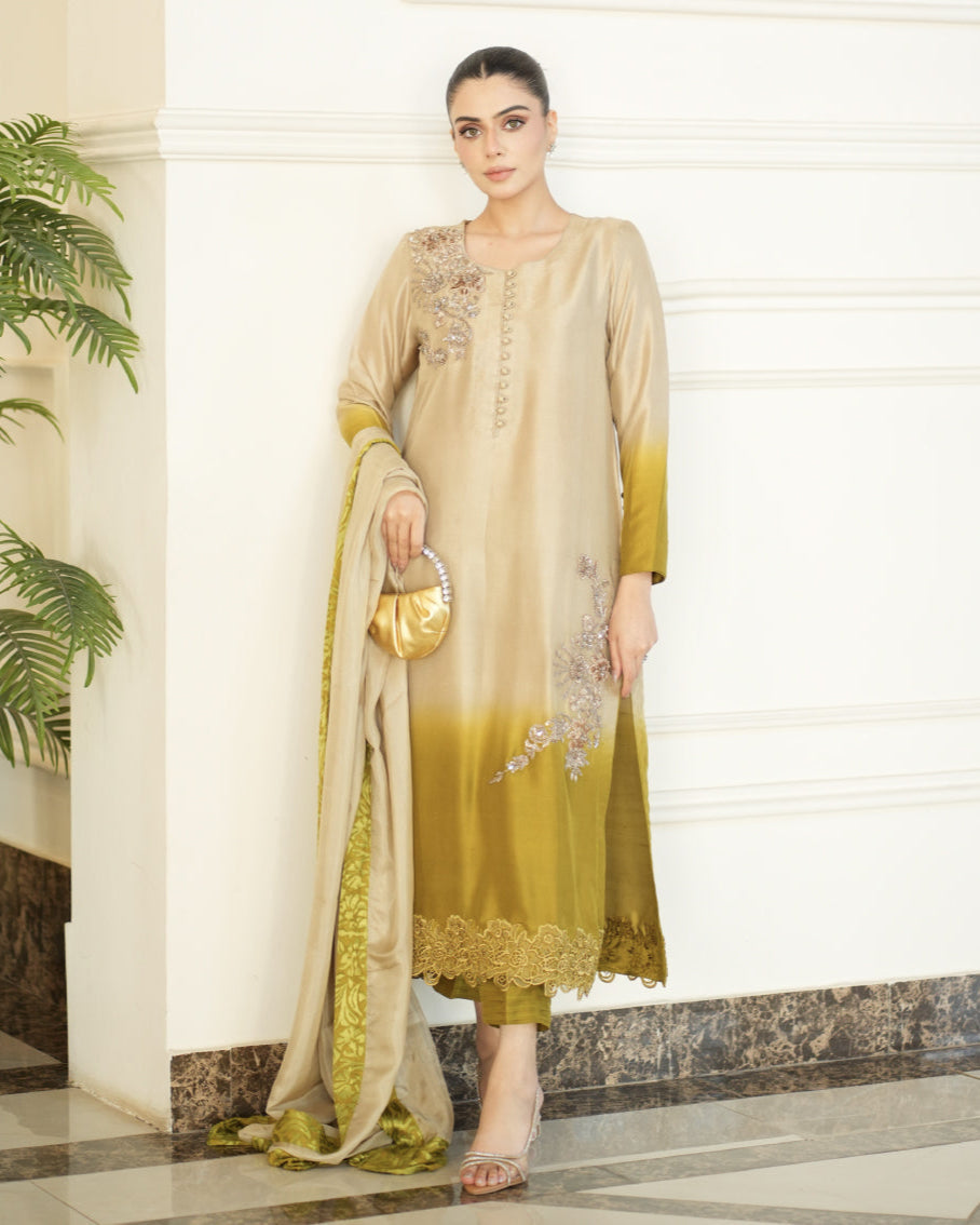 Woman in a beige and green traditional outfit standing indoors with plants and a white wall in the background.