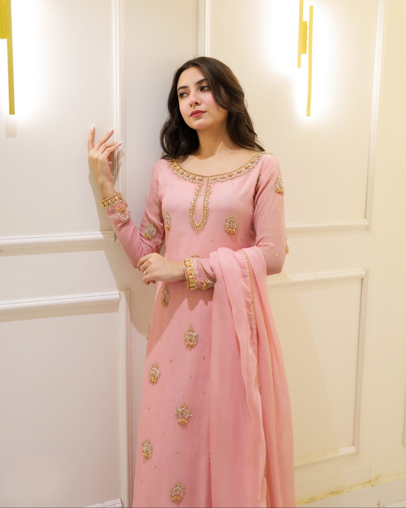 Woman in a pink traditional outfit with gold embroidery standing against a light-colored wall.