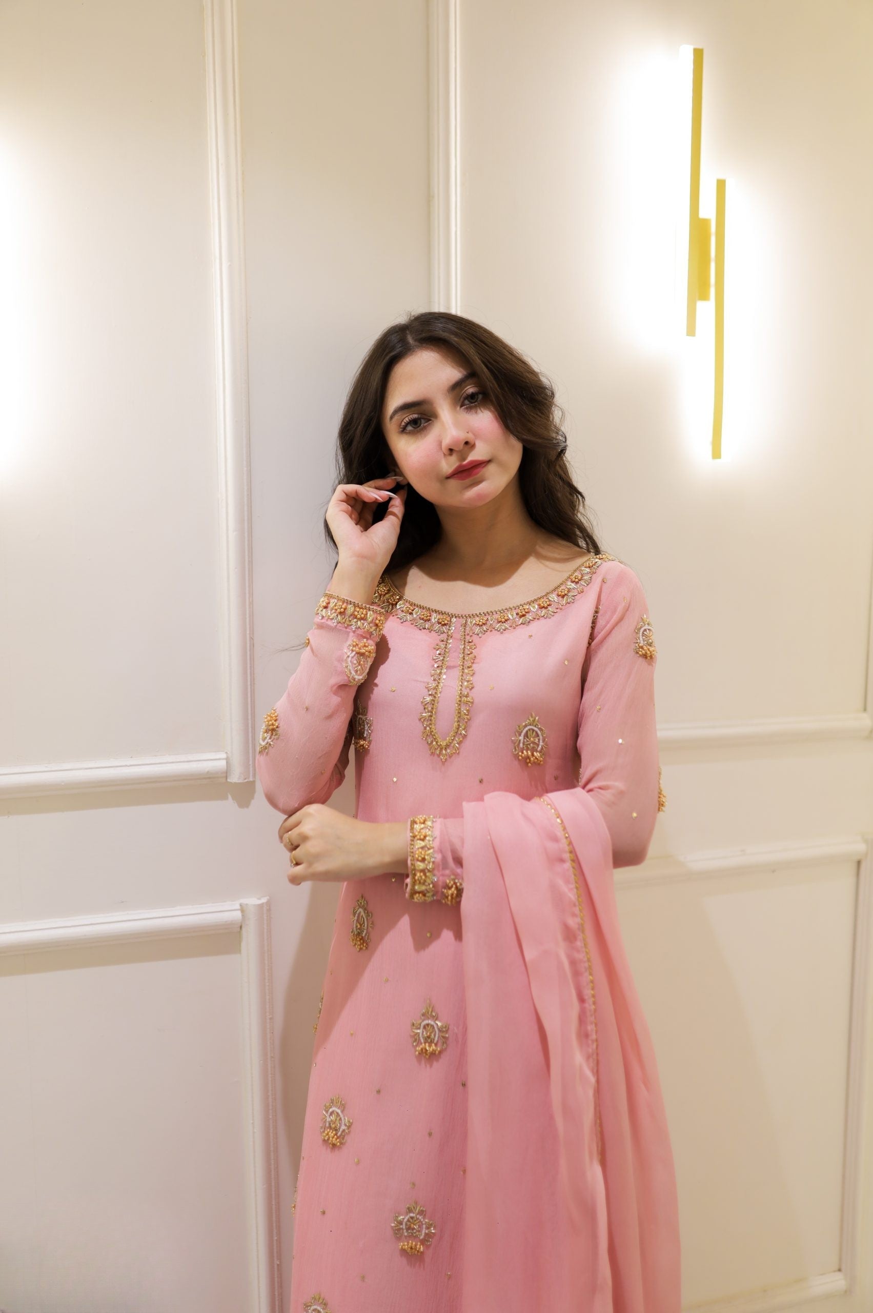 Woman in a pink traditional outfit with gold embroidery against a white wall.
